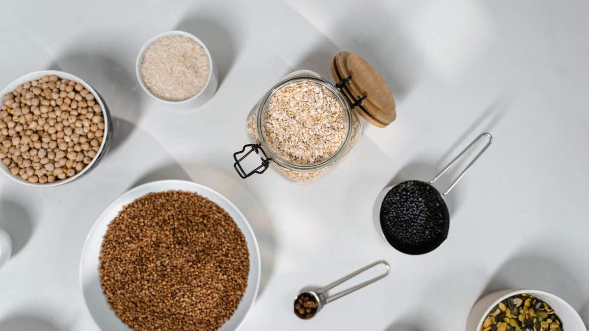 Top view of various grains and seeds in bowls and jars on a white surface.